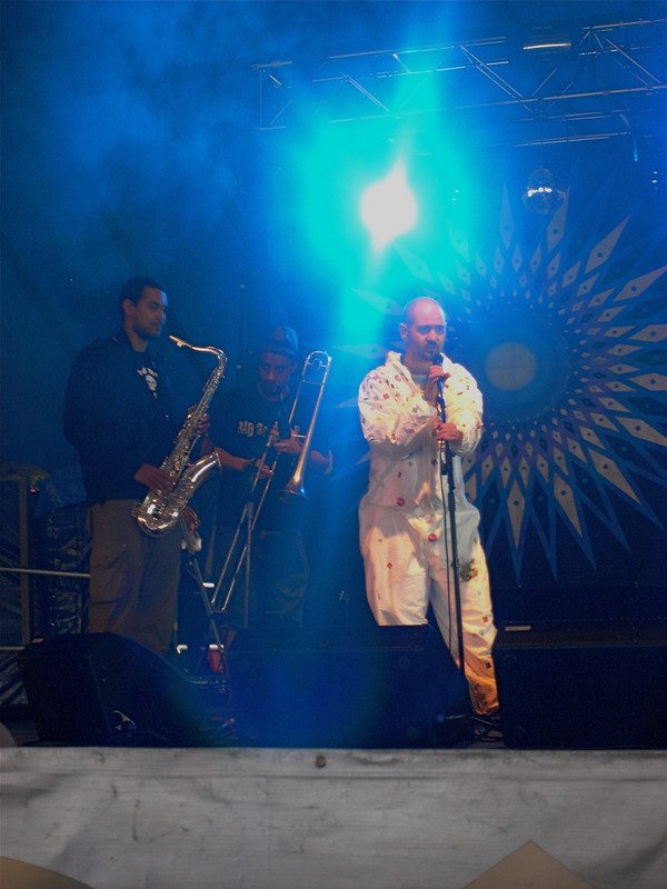 Performer in white overalls on a blue-lit festival stage with sax and trombone, Glastonbury