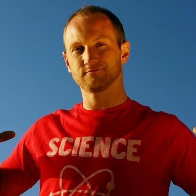 Portrait of Baba Brinkman in a red SCIENCE t-shirt against a blue sky, pointing at himself with both thumbs